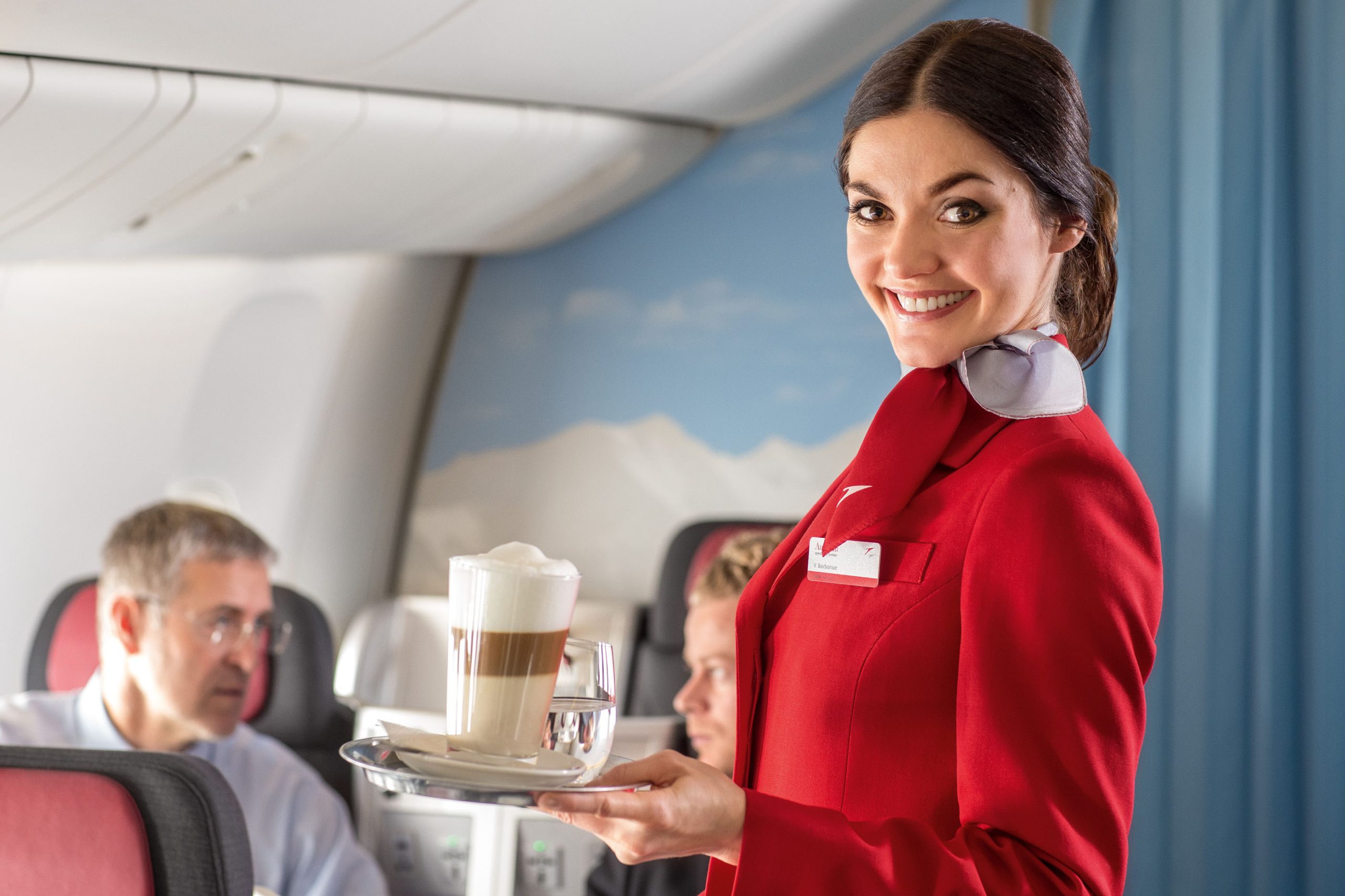 Photo of a smiling woman in a red uniform carrying coffee to seated airplane passengers.