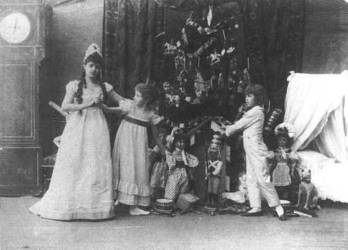 A group of children in festive dresses and hats joyfully gather around a beautifully decorated Christmas tree.