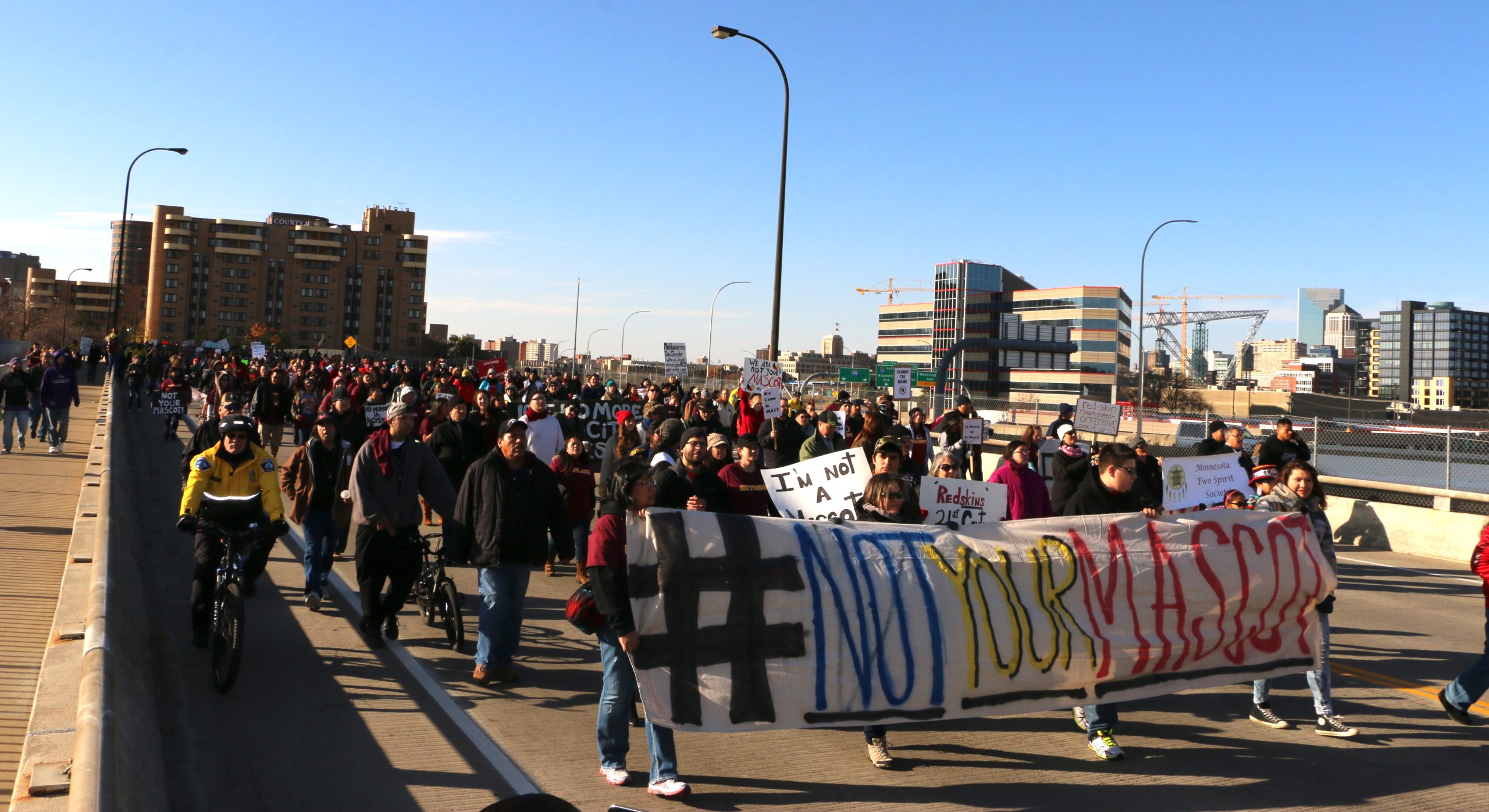 March over an urban bridge. People in front are holding sign saying "Not your mascot". Photo captures hundreds of people in the march.